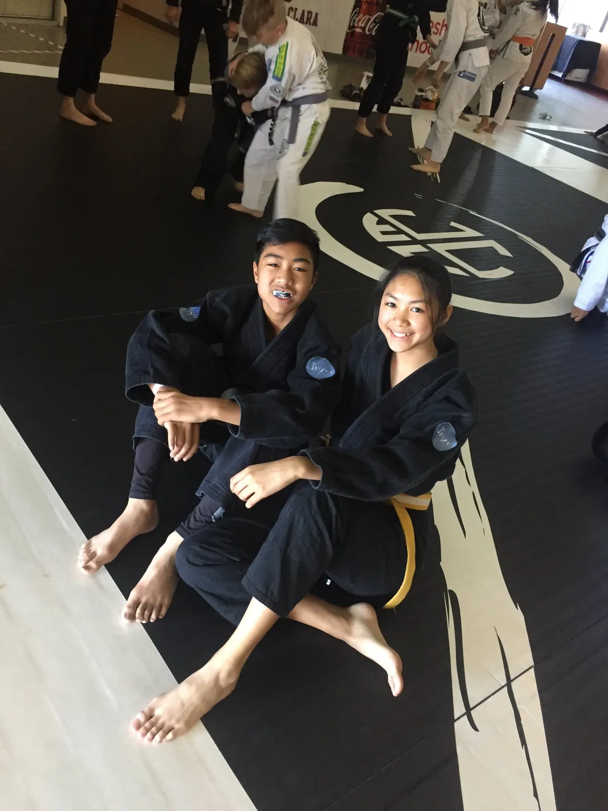 Two smiling Jiyu teens in black gis sitting on the mat at a tournament